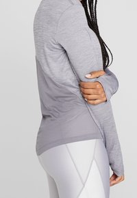 Person med flätat hår som bär en ljusgrå långärmad träningströja med ett nätpanel bak och matchande leggings, med armarna korsade över kroppen.