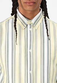 Camicia a maniche lunghe a strisce gialle e bianche con dettagli grigi, dotata di colletto e bottoni frontali. Tessuto a trama fitta con linee verticali.