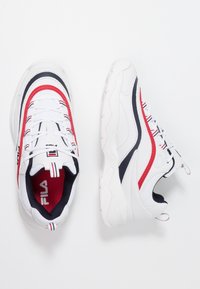 White sneakers with red and navy accents, mesh and synthetic material, rounded toe, textured sole, and laces. Fila logo on the tongue.