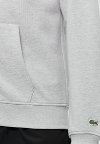 Sweat-shirt gris clair avec ourlet et poignets côtelés, poche kangourou et petit logo crocodile vert sur la manche près du poignet.