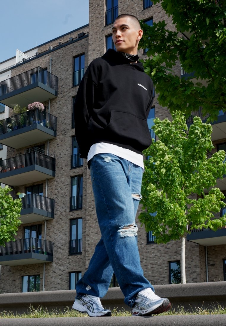 Haut à capuche noir avec logo, sous-couche blanche, jean bleu délavé, baskets grises et blanches, marchant près d'un bâtiment moderne avec de la verdure.