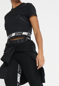 Svart kortärmad träningströja med mönstrad textur, försedd med en logoband vid nederkanten. Matchas med högmidjade svarta leggings, även de med logga.