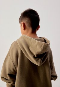 Chłopiec z krótkimi ciemnymi włosami, ubrany w taupe'ową bluzę z kapturem, widoczny od tyłu na tle jednolitego jasnego tła.