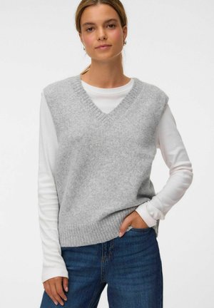 Jeune femme portant un gilet en maille sans manches gris clair sur une chemise blanche à manches longues et un jean bleu, debout avec une main dans la poche.