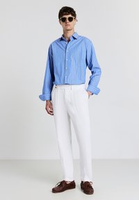 Uomo con occhiali da sole che indossa una camicia a righe blu, pantaloni bianchi testurizzati e scarpe da barca in pelle marrone, in piedi su uno sfondo neutro.