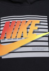 Crna dukserica s kapuljačom koja ima Nike logo u smjelim crvenim i narančastim gradijentima, sivo prugasta pozadina i bijeli tekst s informacijama o zaštitnom znaku.