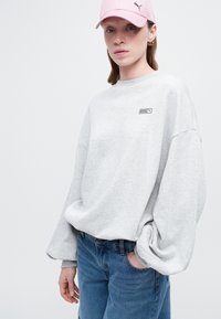Mujer joven vistiendo un sudadera gris claro holgada, jeans azules, una gorra rosa de Puma y pendientes de aro dorados, frente a un fondo blanco liso.
