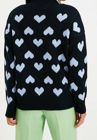 Schwarzer, gestrickter Pullover mit hellblauen Herzmustern, geripptem Kragen und Saum, lockere Passform, kombiniert mit hellgrünen Hosen.