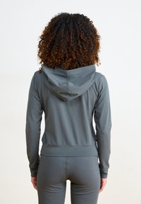 Sudadera con capucha gris de diseño ajustado, con bolsillo tipo canguro y capucha. Tejido suave con textura lisa, combinada con pantalones a juego. Detalles de costura simples.