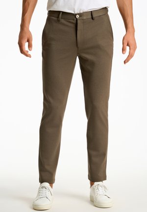 Pantalons marron skinny avec une texture lisse, présentant une fermeture zippée, un bouton de fermeture et des poches avant, assortis à des baskets blanches.