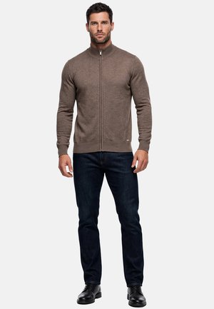 Uomo con maglione marrone con zip, jeans blu scuro e scarpe nere, in piedi davanti a uno sfondo chiaro e uniforme.
