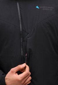 Mână care ține fermoarul unei jachete negre Klättermusen Gondul 2.0, evidențiind textura materialului și sigla de pe piept.