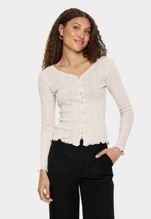 Woman with curly hair wearing a cream buttoned cardigan with lace patterns and black pants, posing with one hand in pocket and slight smile.