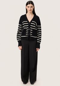 Svart cardigan med vita horisontella ränder, utrustad med V-ringning, fyra knappar och två framfickor. Tillverkad av ribbat tyg med en mjuk textur.