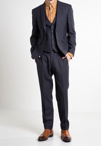 Dreiteiliger Anzug in Dunkelblau mit kariertem Muster, bestehend aus einer taillierten Jacke, einer Weste und maßgeschneiderten Hosen, kombiniert mit einem braunen Hemd und Lederschuhen.