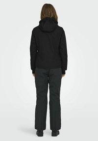 Veste à capuche noire au design ajusté, associée à un pantalon vert foncé avec des poignets zippés, le tout fabriqué à partir de matériaux résistants aux intempéries.
