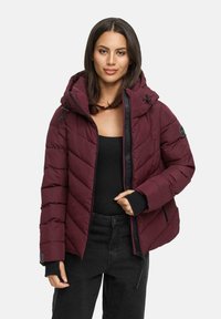 Vrouw in een bordeauxgekleurde gewatteerde pufferjack met capuchon, gedragen over een zwart topje en zwarte broek, staande tegen een lichte achtergrond.