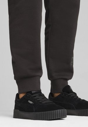 Black suede sneakers with a thick rubber sole and textured detailing, paired with dark fitted sweatpants featuring a logo accent.