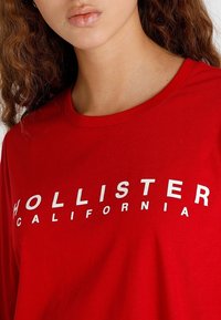 Röd bomull T-shirt med rund hals, långa ärmar och vit text "HOLLISTER CALIFORNIA" tryckt över framsidan.