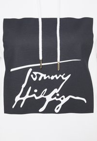 Sudadera blanca con un gran gráfico rectangular negro que exhibe el nombre "Tommy Hilfiger" en texto cursivo blanco, acentuado con detalles dorados.