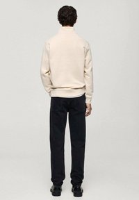Hellbeiger Pullover mit hohem Kragen und gerippten Bündchen, kombiniert mit schwarzen geraden Jeans und schwarzen Schuhen, von hinten gezeigt.