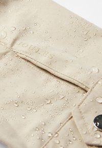 Beige waterproof fabric with water droplets on the surface and visible stitched seam and metallic snap button.