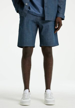 Man wearing mid-thigh dark denim shorts, matching denim jacket, blue shirt, and white sneakers standing against white background.