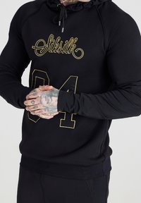 Persona muscolosa che indossa una felpa nera con il testo dorato "Siksilk 84", mani congiunte che mostrano tatuaggi su dita e polso.