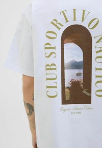 Bela kokvilnas T-kreklu ar brūnu grafiku, kurā attēlots arku logs ar ainavu un zaļo tekstu "CLUB SPORTIVO NAUTICO".