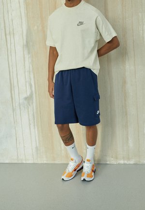 Man draagt een beige Nike T-shirt, marineblauwe Nike korte broek met zak, witte Nike sokken en oranje-witte Nike sneakers, staand binnen.