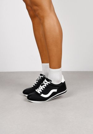Black leather sneakers with white accents, featuring white laces and a rubber sole, paired with white crew socks.