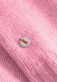 Różowa dzianina z ribbową teksturą, z okrągłą, srebrną metką z czerwoną sylwetką psa przymocowaną czerwoną nitką.