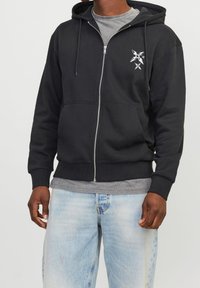 Felpa nera con zip realizzata in cotone, dotata di tasca frontale, cappuccio con lacci e una grafica sul petto sinistro. Indossata con jeans azzurri chiari.