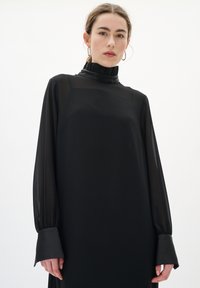 Černé dlouhé šaty s vysokým volánkovým límcem, průhlednou látkou na rukávech a přiléhavými manžetami. Hladká textura, minimalistický design.