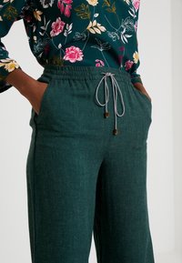 Tealgröna lätta byxor med elastisk midja och dragsko; sidofickor med subtil textur. Kombination med blommönstrad blus.