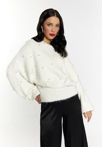 Pulover alb, tricotat, cu croială lejeră și mâneci tip balon, împodobit cu perle false uniform distribuite. Asortat cu pantaloni negri largi.