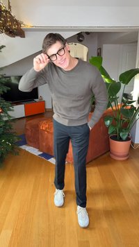 Grijze geribbelde trui, zwarte broek en witte sneakers. Staand op een houten vloer met groene planten en een oranje bank op de achtergrond.