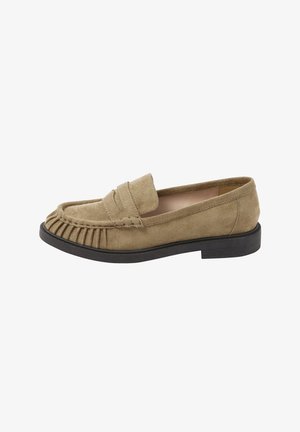 Beige ruskindslæder med folder på tåen, sort sål, lav hæl og slip-on design.