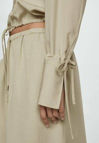 Beige long-sleeve blouse with elastic cuffs and adjustable ties, featuring a relaxed fit and lightweight fabric. Smooth texture, minimal design.