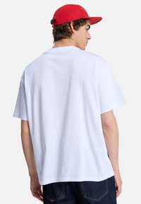 Hvid bomulds t-shirt med korte ærmer og rund halsudskæring, vist fra ryggen. Parret med mørke denim bukser og en rød cap.