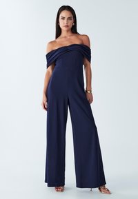 Tamsiai mėlynas peties neslepiantis kombinezonas su klostuota apykakle, aptempta liemene ir plačiomis kelnėmis. Pagamintas iš lygaus, tamprios medžiagos. Auksinės basutės.