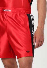 Red satin athletic shorts with black side stripes, elastic waistband, and small black Adidas logo on the lower hem. Smooth texture.
