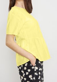 Top peplum giallo brillante a maniche corte realizzato in tessuto morbido, abbinato a pantaloni neri a fantasia con disegni floreali bianchi.