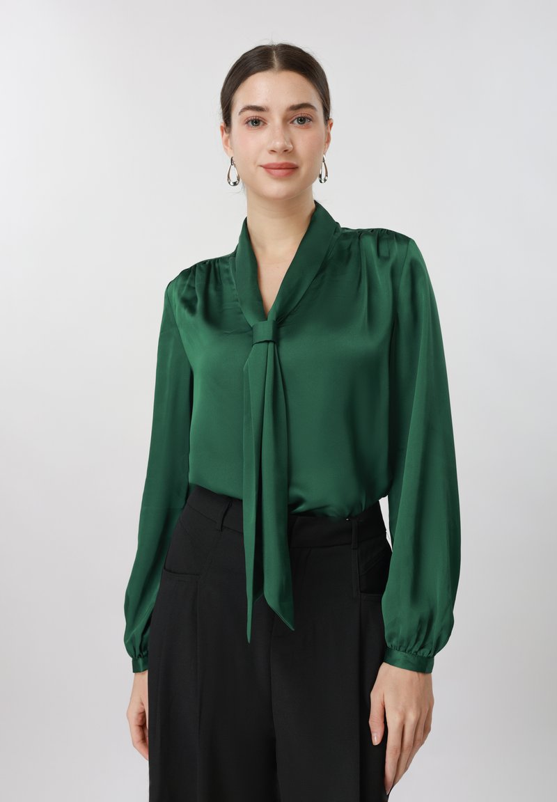 Femme portant un chemisier en satin vert émeraude à manches longues avec un nœud au col, des boucles d'oreilles créoles, et un pantalon noir taille haute, debout devant un fond blanc.