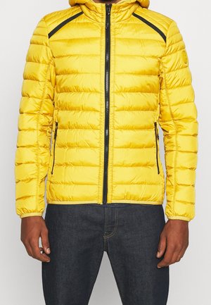 Veste matelassée jaune à capuche avec fermeture éclair noire et détails de poche, portée avec un jean bleu foncé, visible du cou aux hanches.