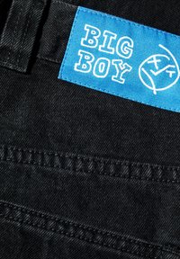 Los pantalones de mezclilla negros presentan una etiqueta de tela azul con texto blanco que dice "BIG BOY". La exhibición incluye material texturizado y detalles de costura.