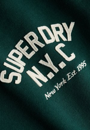 Donkergroene stof met witte geborduurde tekst "SUPERDRY N.Y.C New York Est. 1995" in vetgedrukte en cursieve letters.