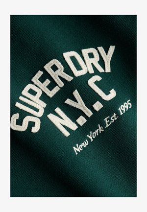 Dark green fabric with white embroidered text "SUPERDRY N.Y.C New York Est. 1995" in bold and cursive fonts.
