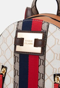 Brun och gräddfärgad ryggsäck med geometriskt mönster, dekorerad med röd och blårandiga detaljer samt guldfärgad hårdvara som pryder River Island-logotypen.