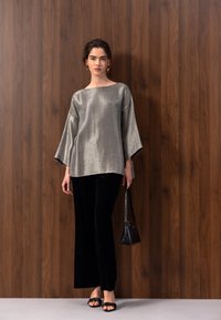 Jeune femme debout contre un mur en bois, portant un haut argenté ample, un pantalon noir à jambes larges, des sandales noires et tenant un petit sac à main noir.